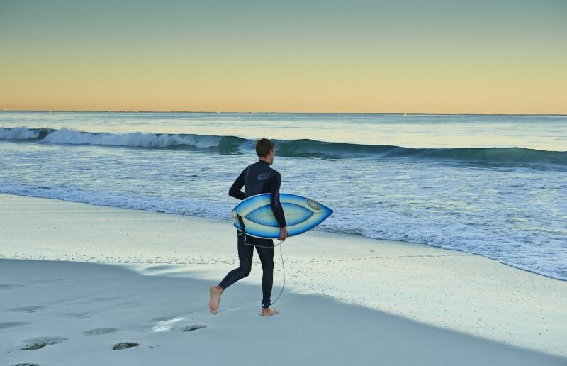 One surfer in wetsuit carrying surfboard run towards the ocean on a sandy beach at sunrise or sunset.