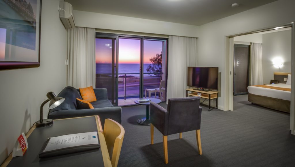 Modern hotel suite with a living area, a separate bedroom, and a balcony overlooking the ocean at sunset.