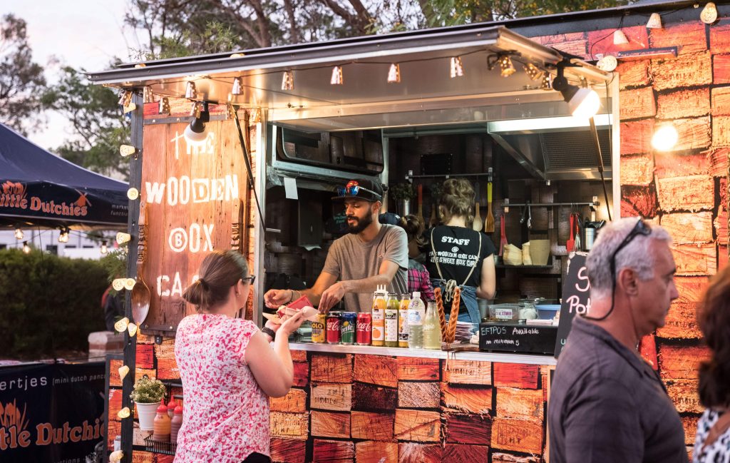A food vendor at a wooden-themed food truck serves a customer at an outdoor night market.