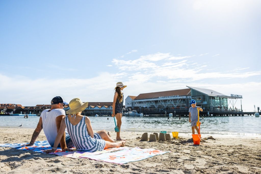 A family relaxes on Hillarys Boat Harbour near calm waters, with children playing in the sand and a marina with a large waterfront building in the background.