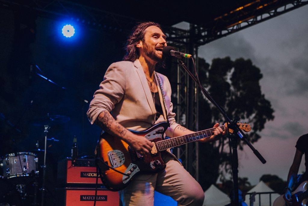 A male musician in a beige suit performs on stage at dusk, singing into a microphone while playing an electric guitar.