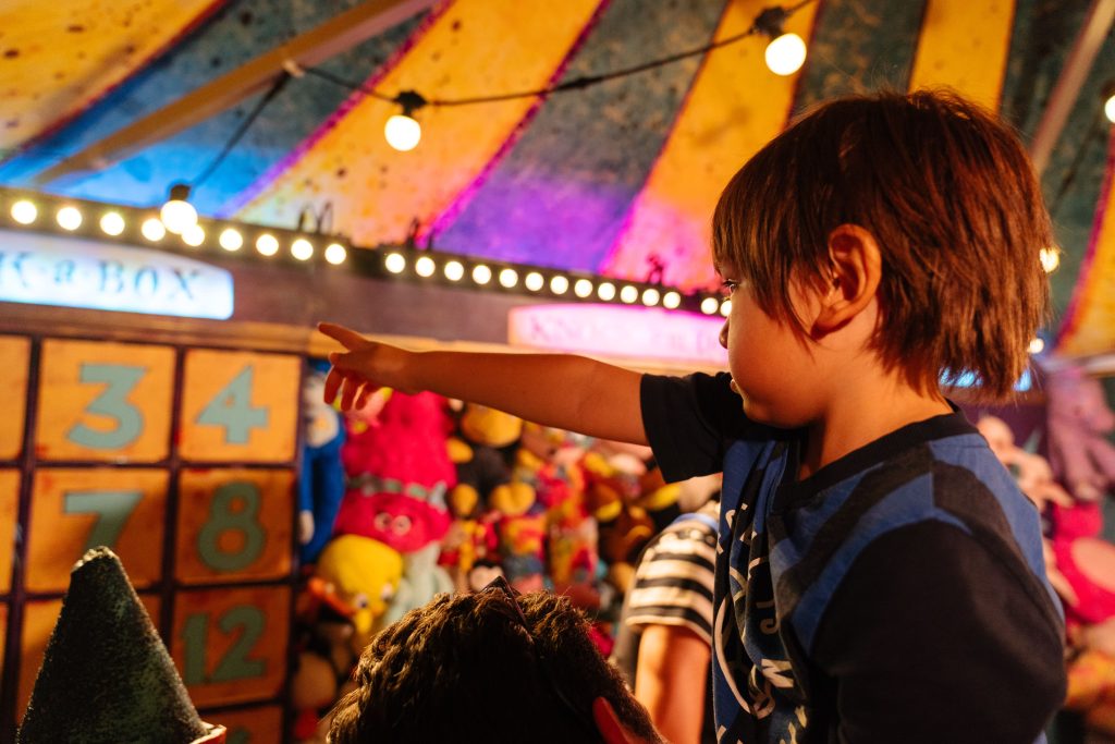 A young child on someone's shoulders excitedly points at a carnival game booth filled with plush toys.