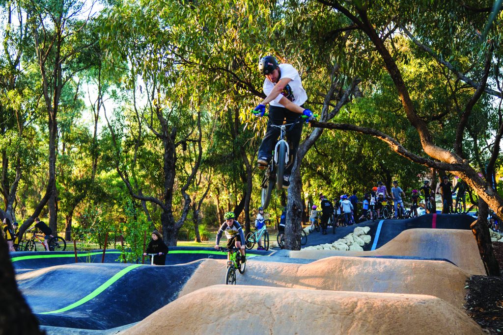A BMX rider mid-air performing a trick at Shepards Bush surrounded by trees, with other cyclists and spectators in the background.
