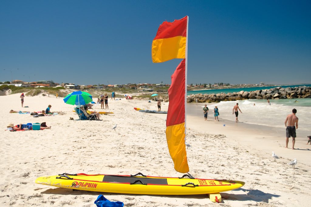 A red and yellow surf lifesaving flag on Sorrento Beach with a rescue board nearby, surrounded by beachgoers and ocean waves.