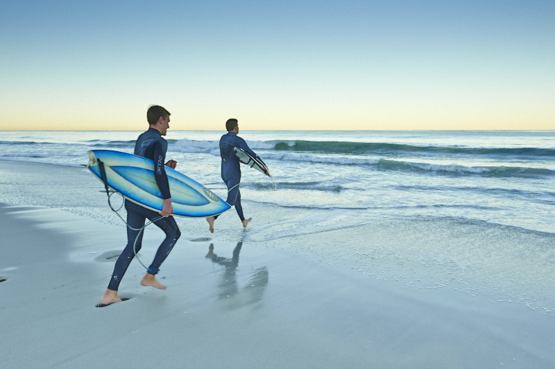 Two surfers in wetsuits carrying surfboards run towards the ocean on a sandy beach at sunrise or sunset.