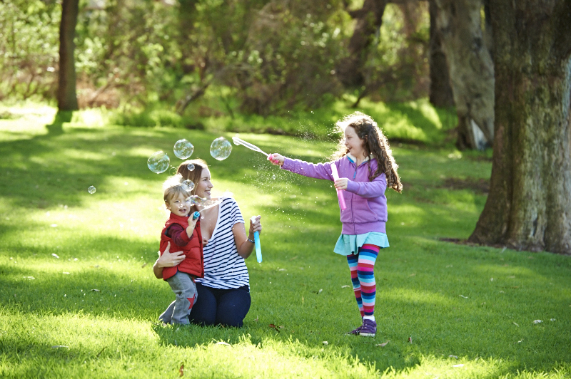 A mother and two children play with bubbles in a sunlit park surrounded by green grass and trees.