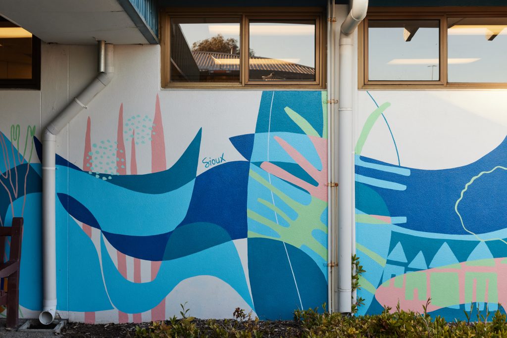 A colorful abstract mural with organic shapes and patterns in shades of blue, green, and pink, painted on an exterior wall. Credit: Sioux Tempestt