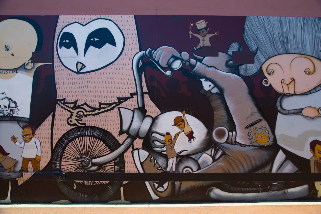 A colorful mural featuring surreal characters, including an owl-faced figure, a motorcyclist, and small animated people interacting. Credit: Stormie Mills